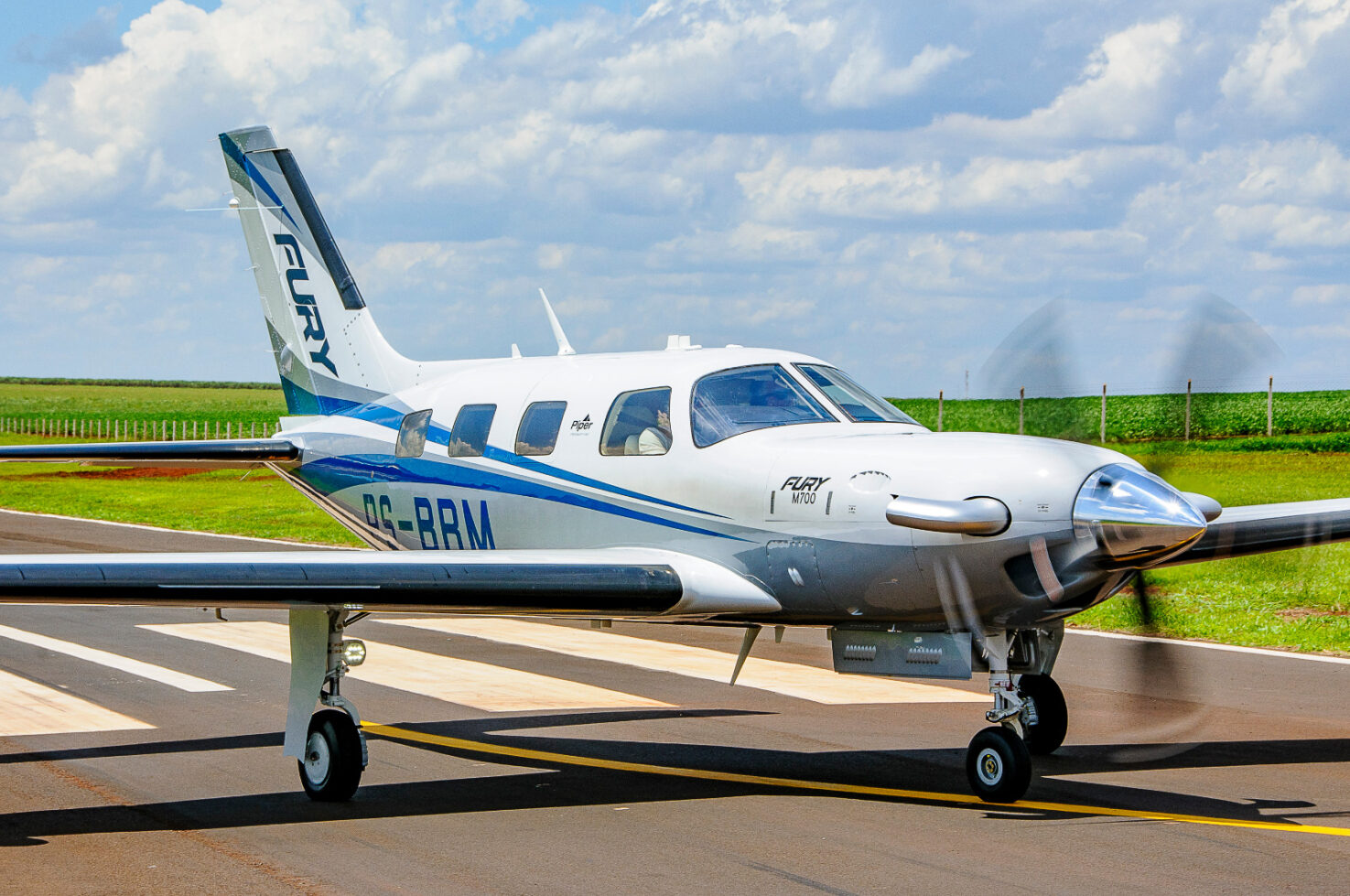 Side view of a Piper M700 Fury taxiing on a runway with propeller spinning and green fields in the background.